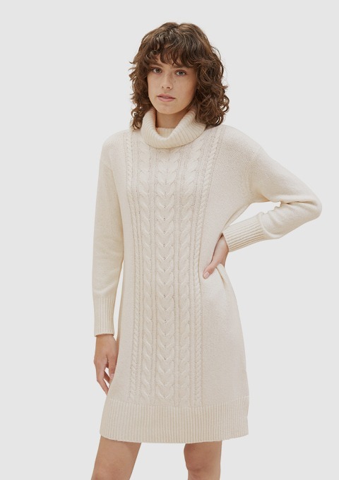 Eine Person trägt ein creamfarbenes Strickkleid mit Rollkragen und Zopfmuster. Eine Person trägt ein creamfarbenes Strickkleid mit Rollkragen und Zopfmuster.