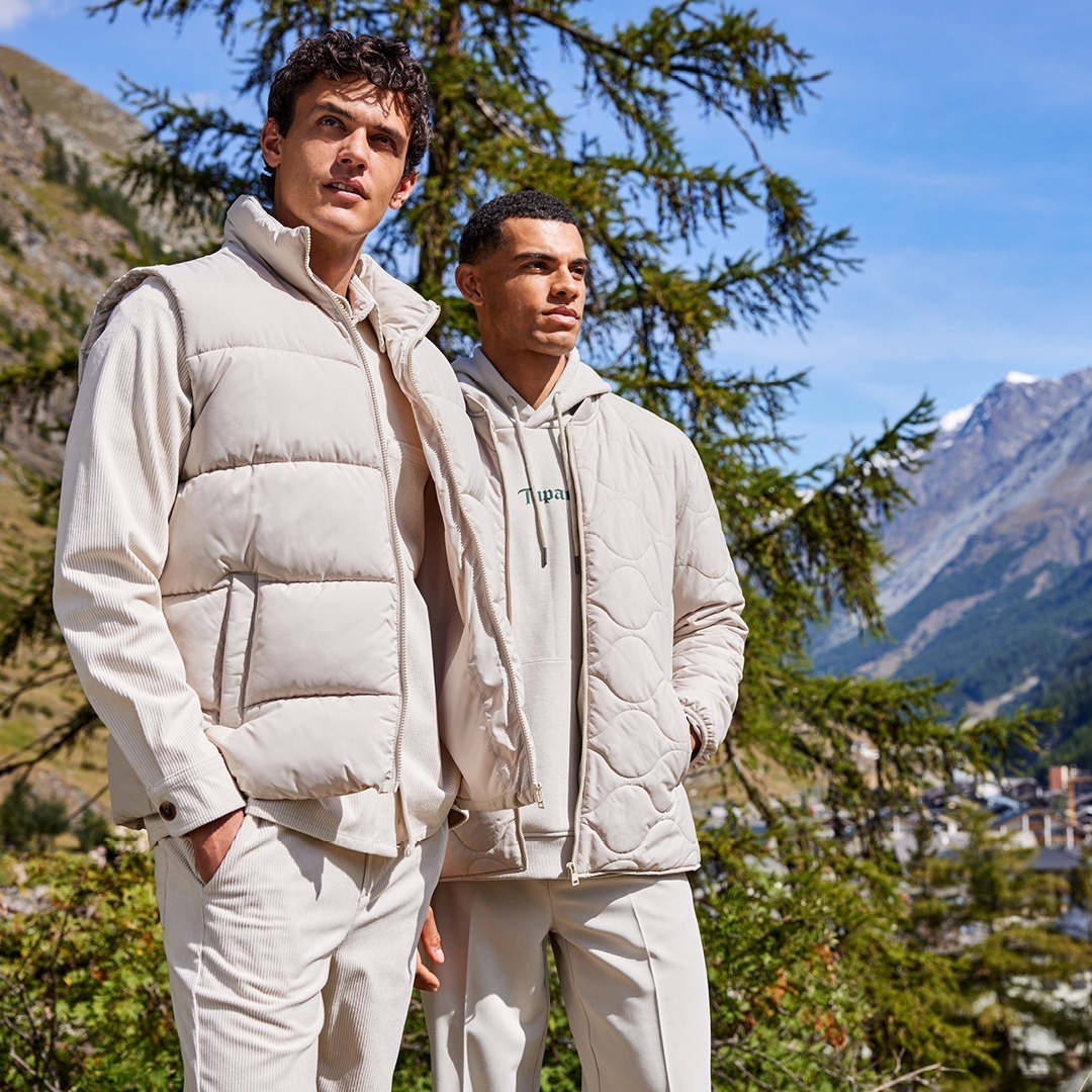 Zwei Männer, die in der Natur stehen, tragen elegante, helle Kleidung. Im Hintergrund sind Berge und Bäume zu sehen. Zwei Männer, die in der Natur stehen, tragen elegante, helle Kleidung. Im Hintergrund sind Berge und Bäume zu sehen.