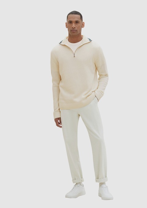 Ein Mann steht in einem cremefarbenen Pullover mit Reißverschluss und weißen Hosen. Er trägt weiße Sneaker. Ein Mann steht in einem cremefarbenen Pullover mit Reißverschluss und weißen Hosen. Er trägt weiße Sneaker.