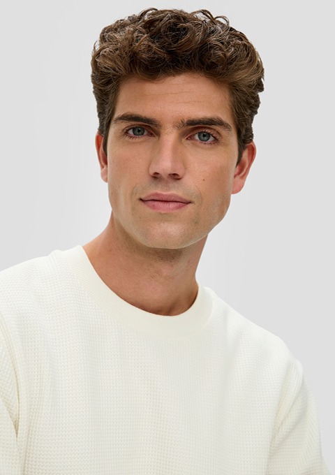 Ein junger Mann mit lockigem, braunem Haar trägt ein weißes Sweatshirt. Er schaut direkt in die Kamera mit neutralem Ausdruck. Ein junger Mann mit lockigem, braunem Haar trägt ein weißes Sweatshirt. Er schaut direkt in die Kamera mit neutralem Ausdruck.