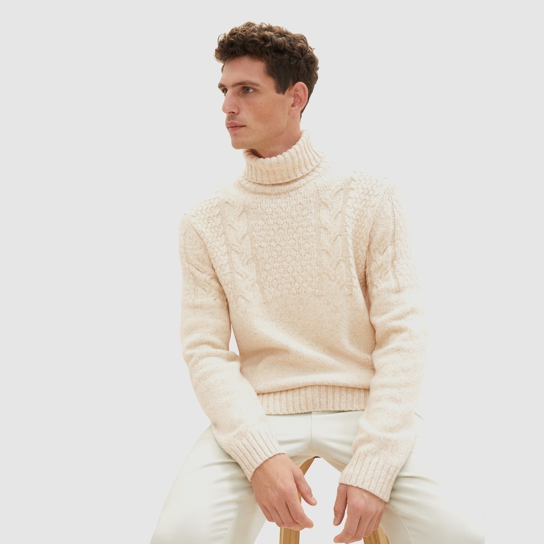 Ein Mann sitzt auf einem Hocker und trägt einen hellen, dick gestrickten Rollkragenpullover. Ein Mann sitzt auf einem Hocker und trägt einen hellen, dick gestrickten Rollkragenpullover.