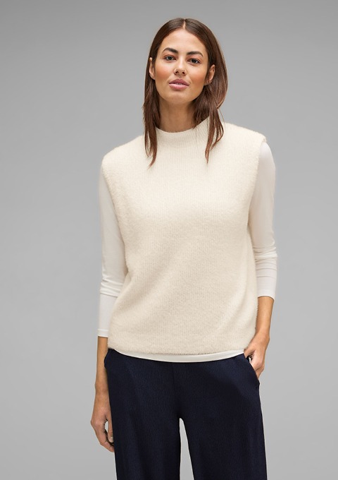 Eine Frau trägt einen weißen, ärmellosen Pullover über einem langen, weißen Shirt und dunkelblauen Hosen. Eine Frau trägt einen weißen, ärmellosen Pullover über einem langen, weißen Shirt und dunkelblauen Hosen.