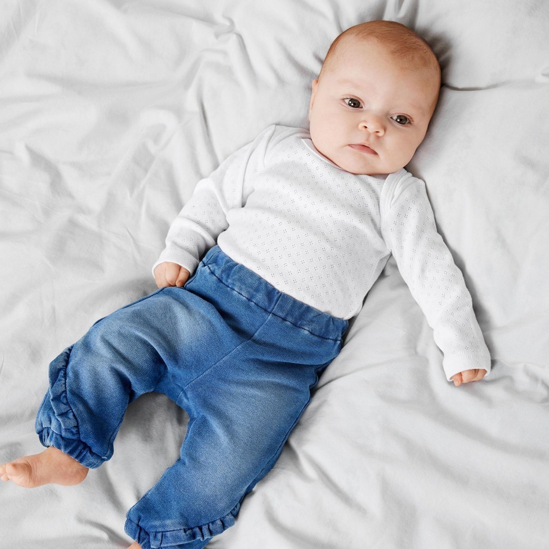 Ein Baby in einem weißen Langarmshirt und blauen Hose liegt auf einem Bett mit weißen Laken. Ein Baby in einem weißen Langarmshirt und blauen Hose liegt auf einem Bett mit weißen Laken.