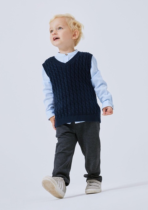 Ein kleiner Junge mit blondem Haar trägt einen blauen gestrickten Pullover, ein hellblaues Hemd und graue Hosen. Er schaut lächelnd nach oben. Ein kleiner Junge mit blondem Haar trägt einen blauen gestrickten Pullover, ein hellblaues Hemd und graue Hosen. Er schaut lächelnd nach oben.