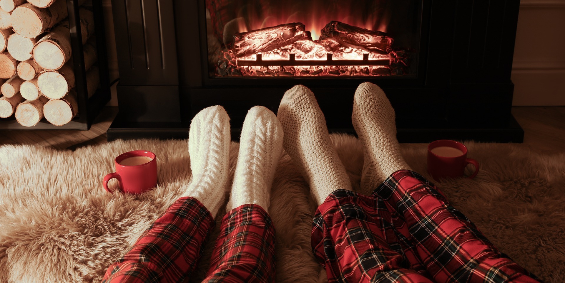 Zwei Personen in gemütlichen Socken und Schlafanzügen entspannen vor einem Kamin. Neben ihnen stehen rote Tassen mit Getränken auf einem weichen Teppich.