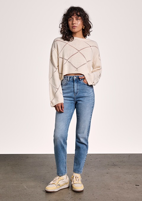 Eine Person trägt einen beigen, gestrickten Pullover und blaue Jeans, kombiniert mit weißen Sneakers.