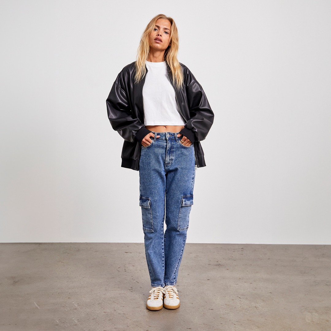 Eine Person trägt eine schwarze Lederjacke, ein weißes Crop-Top und Jeans mit Seitentaschen. Sie steht auf einem neutralen Hintergrund.