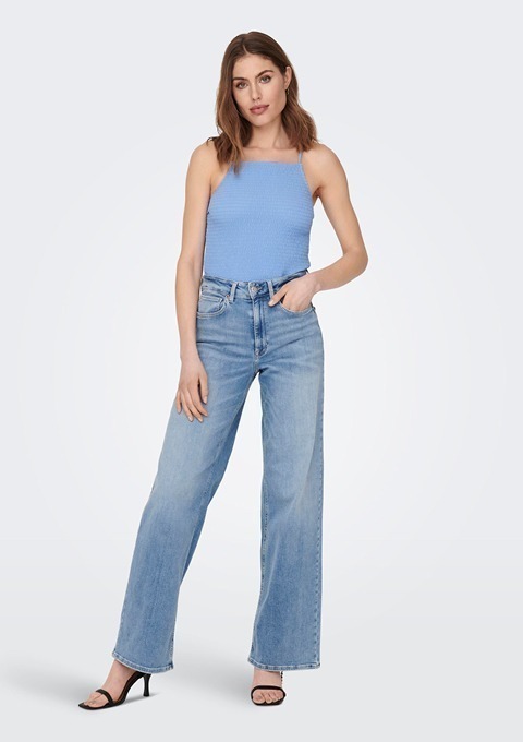 Eine Frau steht in einem blauen Oberteil und weißen Jeans. Sie trägt schwarze Sandalen.