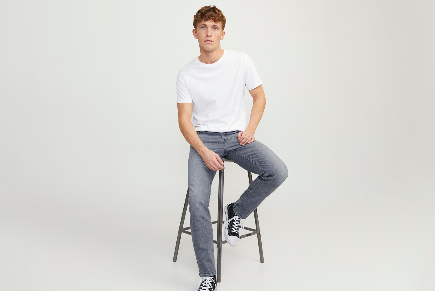 Ein junger Mann sitzt auf einem Hocker, trägt ein weißes T-Shirt und graue Jeans.