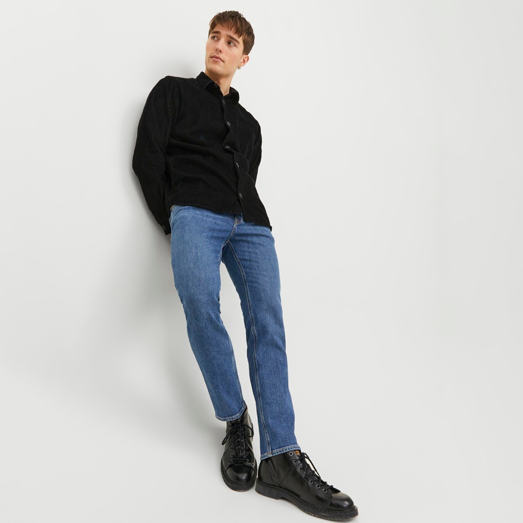 Ein junger Mann steht gegen eine Wand geneigt, trägt ein schwarzes Hemd, blaue Jeans und schwarze Stiefel.