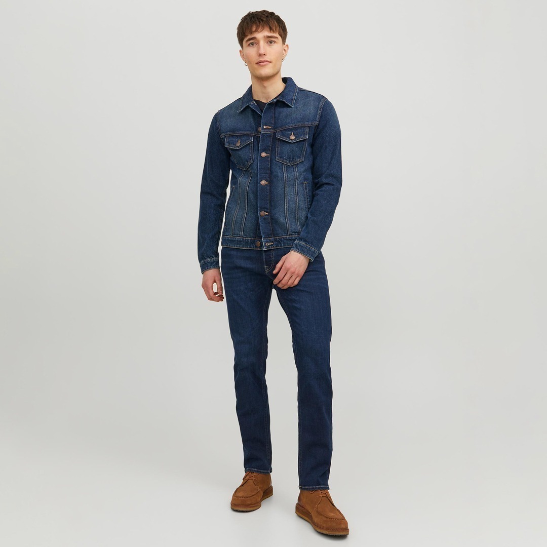 Ein junger Mann in einer blauen Jeansjacke, blauem Jeans und braunen Schuhen steht vor einem hellen Hintergrund.