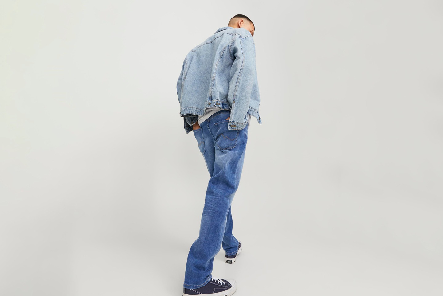 Ein Mann trägt eine Jeansjacke und blaue Jeans. Er steht gedreht, sodass sein Rücken zu sehen ist.