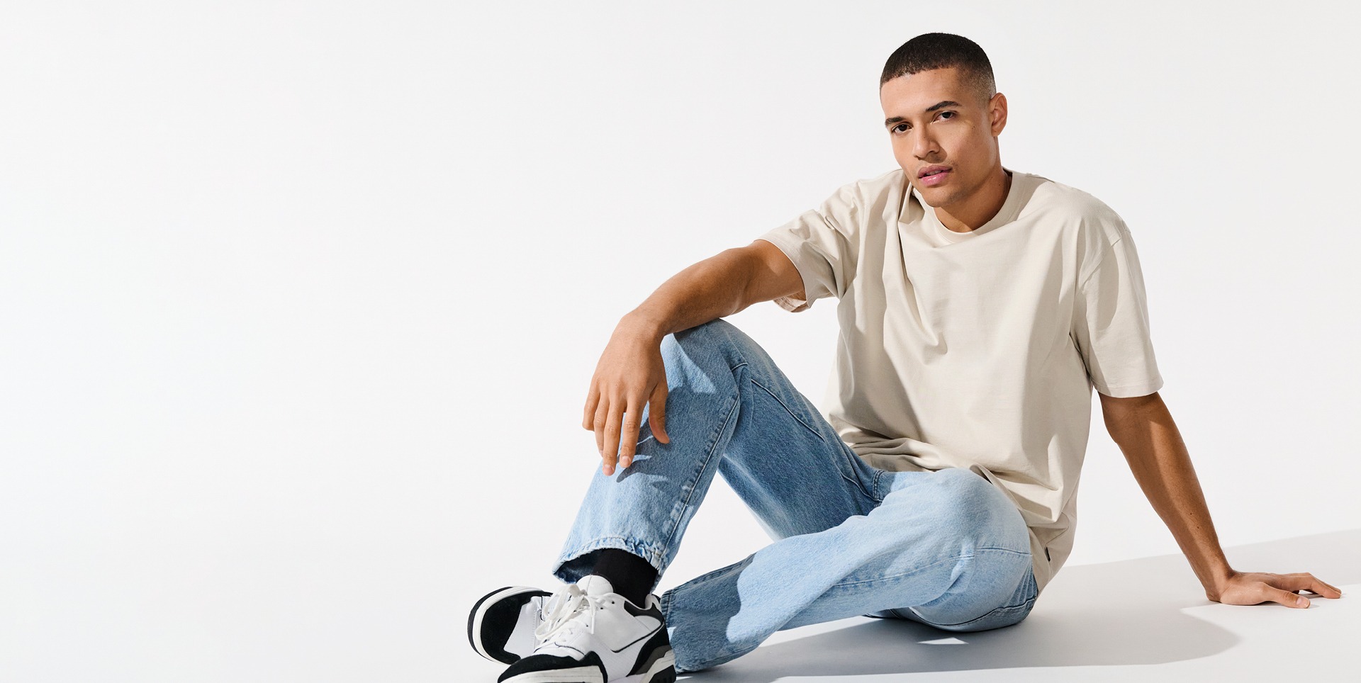 Ein junger Mann sitzt auf dem Boden, trägt ein beiges T-Shirt und blaue Jeans, mit schwarzen und weißen Sneakers. Der Hintergrund ist weiß. Ein junger Mann sitzt auf dem Boden, trägt ein beiges T-Shirt und blaue Jeans, mit schwarzen und weißen Sneakers. Der Hintergrund ist weiß.