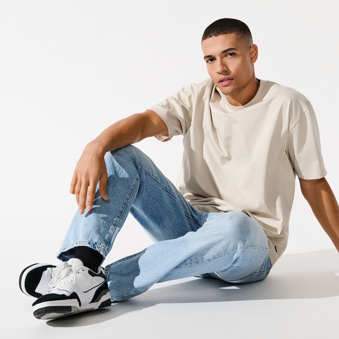Ein Mann sitzt auf dem Boden, trägt ein beiges T-Shirt und hellblaue Jeans, kombiniert mit weißen Sneakern. Ein Mann sitzt auf dem Boden, trägt ein beiges T-Shirt und hellblaue Jeans, kombiniert mit weißen Sneakern.
