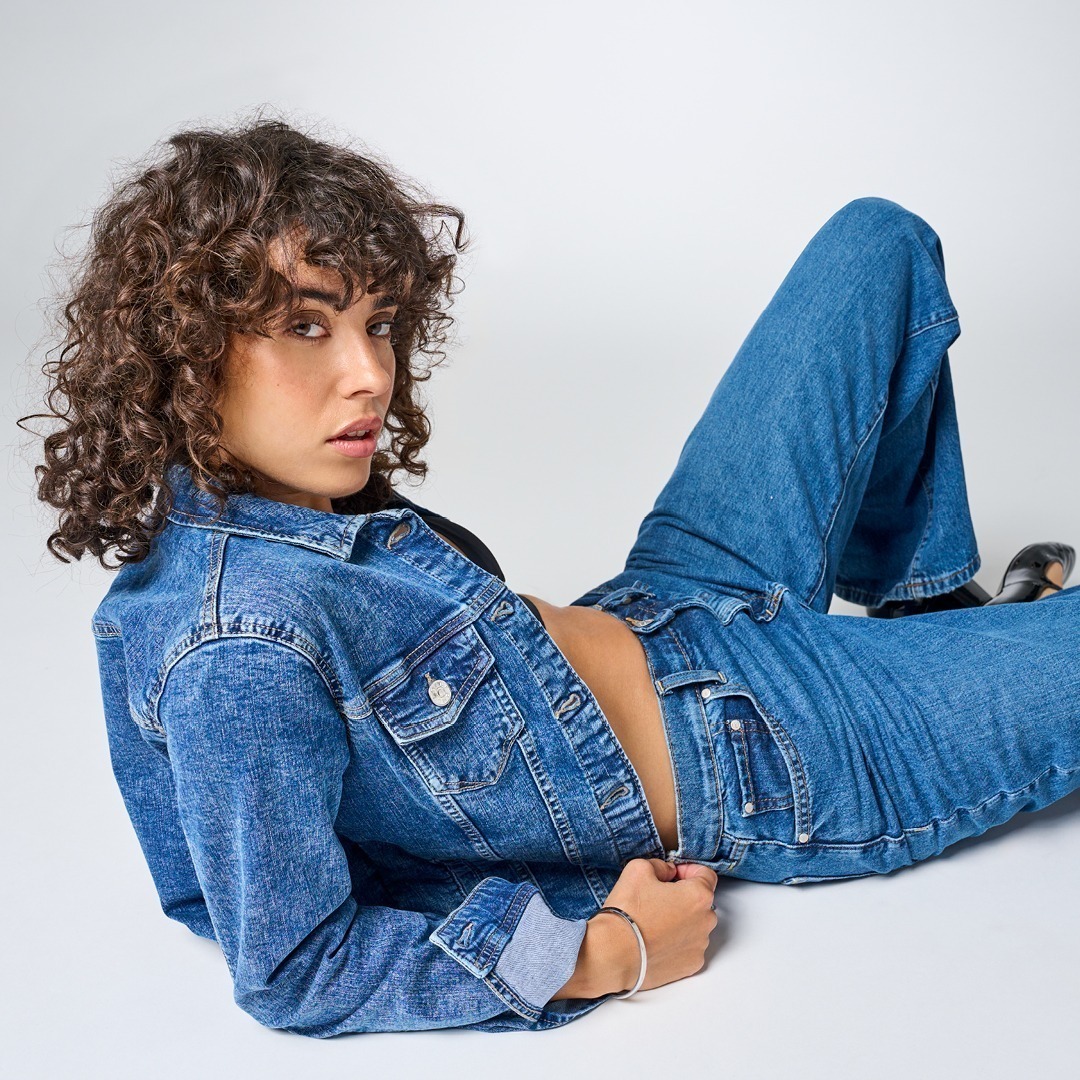 Eine Person mit lockigem Haar trägt eine Jeansjacke und Jeans in derselben Farbtönung. Sie sitzt lässig auf dem Boden und schaut zur Seite. Eine Person mit lockigem Haar trägt eine Jeansjacke und Jeans in derselben Farbtönung. Sie sitzt lässig auf dem Boden und schaut zur Seite.