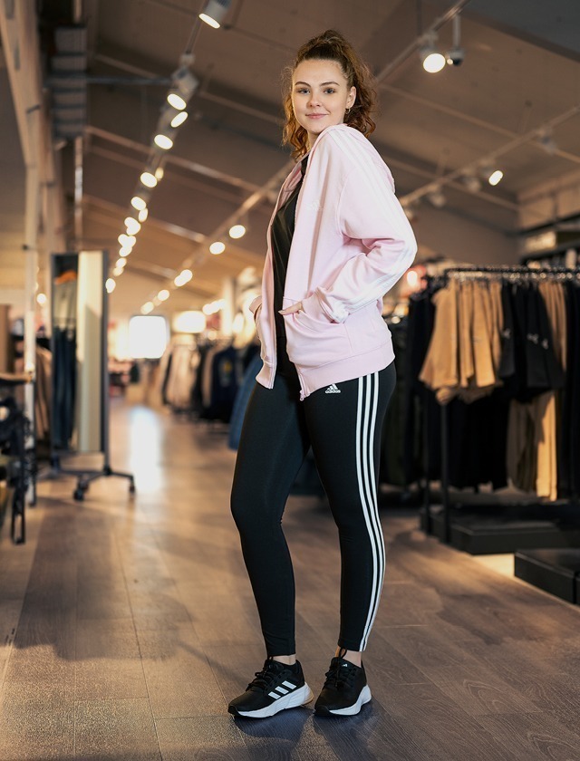 Ein junges Mädchen in sportlicher Kleidung, bestehend aus einer rosa Jacke, schwarzem Oberteil, schwarzen Leggings mit weißen Streifen und Sportschuhen, steht in einem Bekleidungsgeschäft.