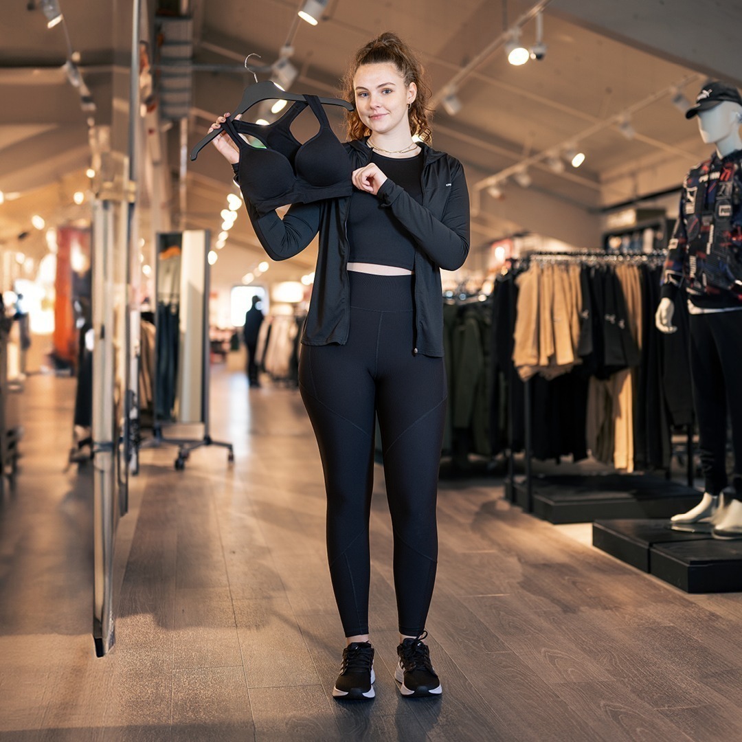 Eine Frau steht in einem Geschäft und hält einen Sport-BH in der Hand. Sie trägt sportive Kleidung und blickt in die Kamera.
