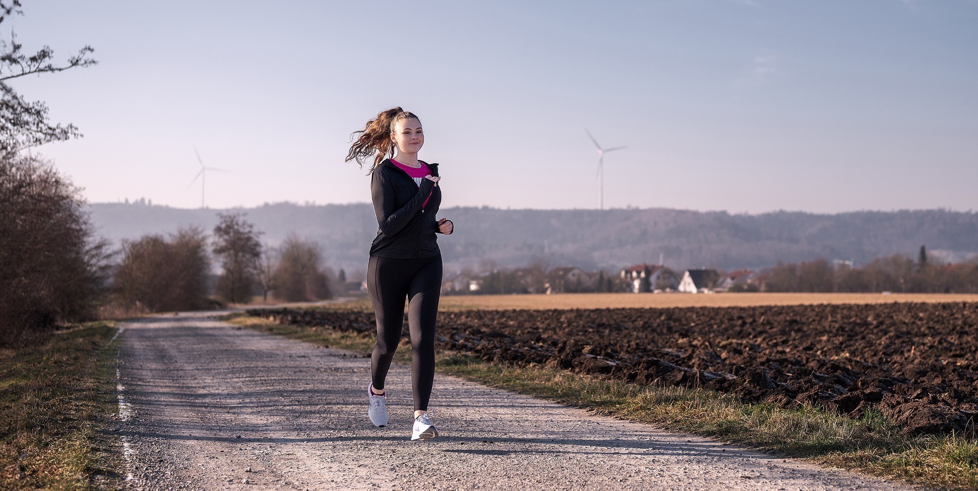 Eine joggende Frau auf einem Weg, umgeben von Feldern und Bäumen, bei klarer Sicht und Windkraftanlagen im Hintergrund.