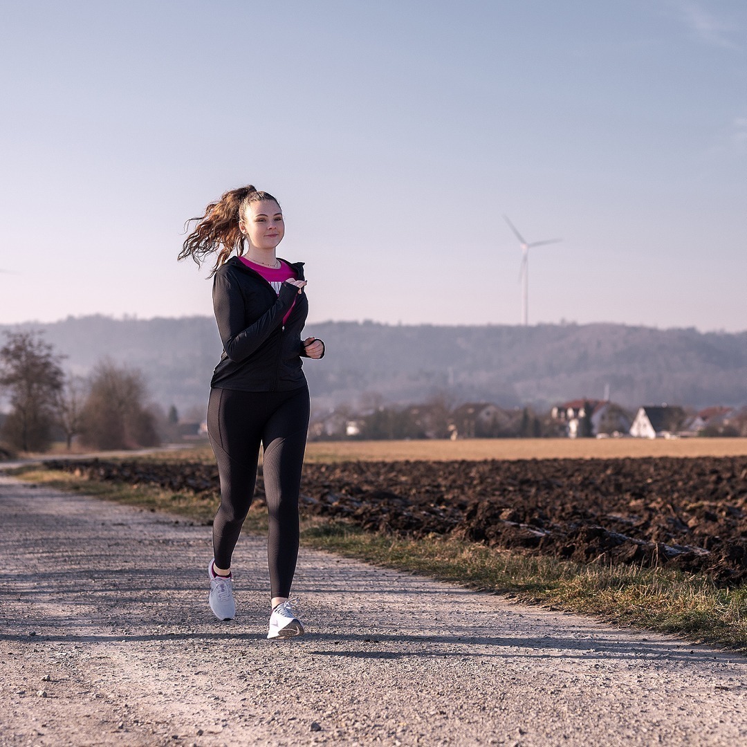 Eine junge Frau läuft auf einem Feldweg, umgeben von einer ländlichen Landschaft und Windrädern im Hintergrund.