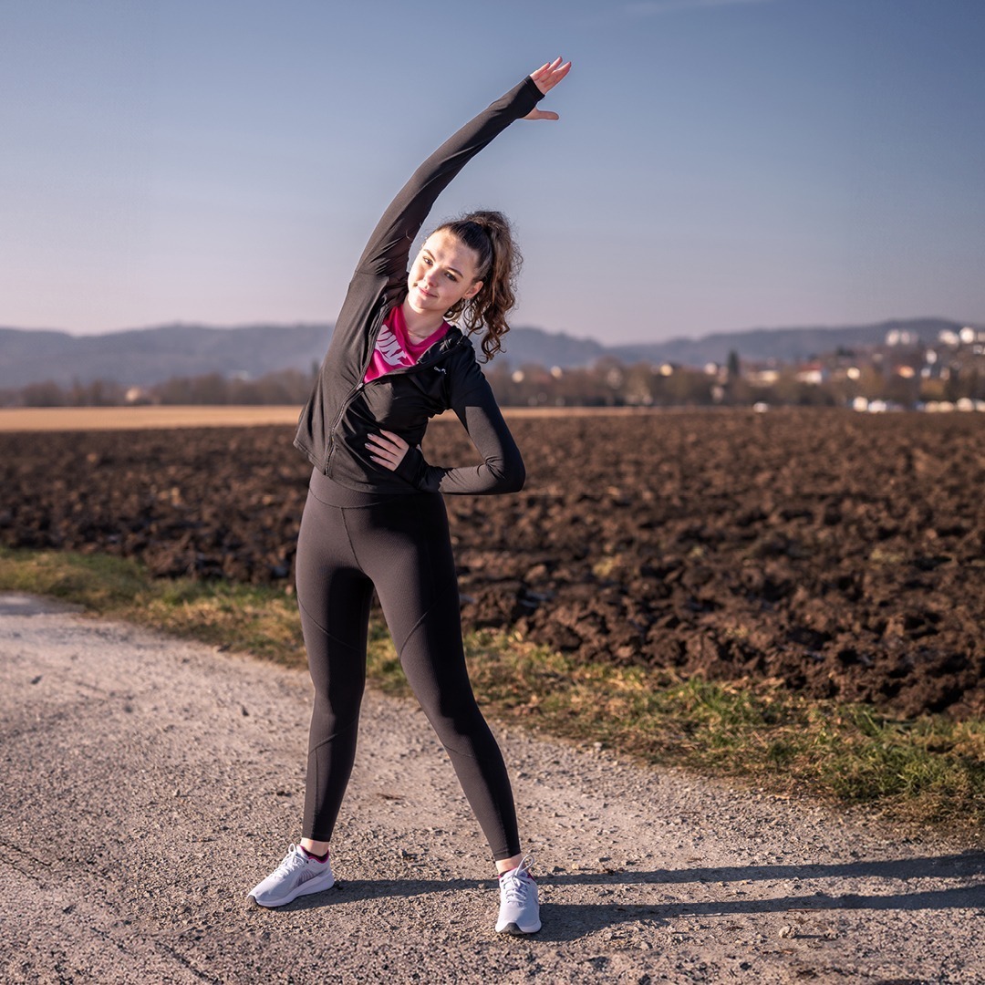 Eine sportliche Frau dehnt sich auf einem Feldweg, trägt schwarze Leggings und ein pinkes Oberteil.