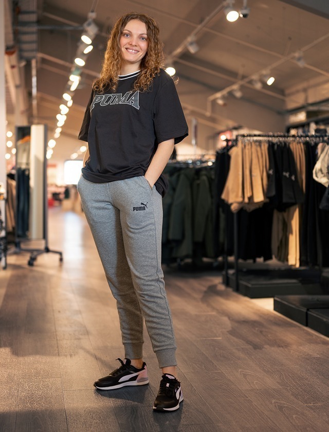 Eine Person steht in einem Bekleidungsgeschäft und trägt ein schwarzes T-Shirt und graue Jogginghosen von Puma, dazu schwarze Sneakers.
