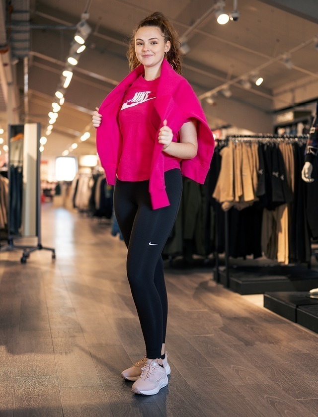 Eine junge Frau steht in einem Geschäft, trägt ein pinkes Nike-Oberteil und schwarze Leggings.