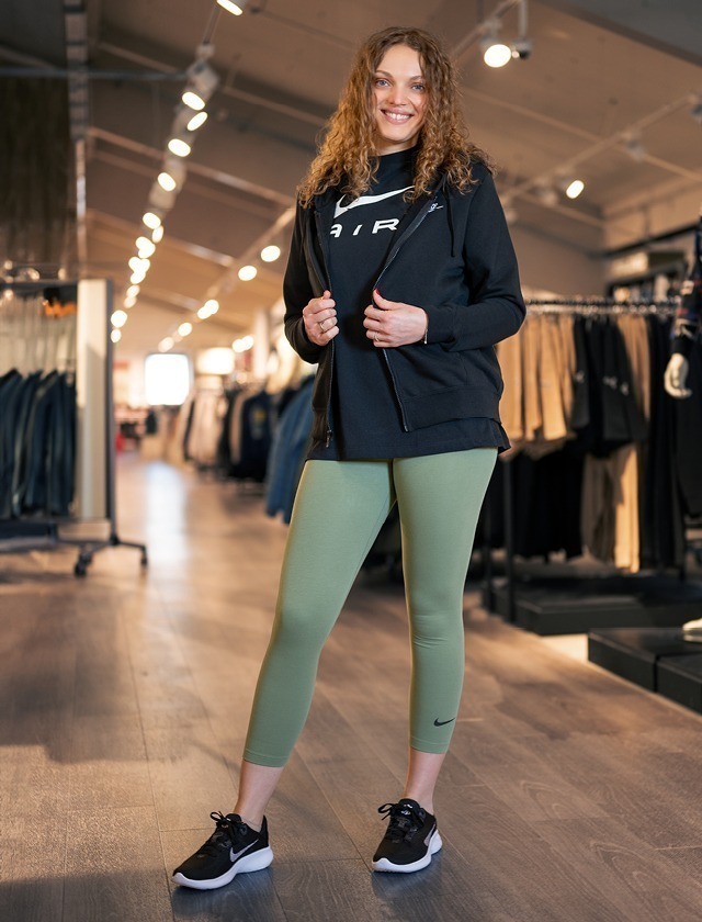 Eine lächelnde Frau in Sportkleidung steht in einem Geschäft. Sie trägt ein schwarzes Oberteil, eine sportliche Jacke und olivgrüne Leggings.