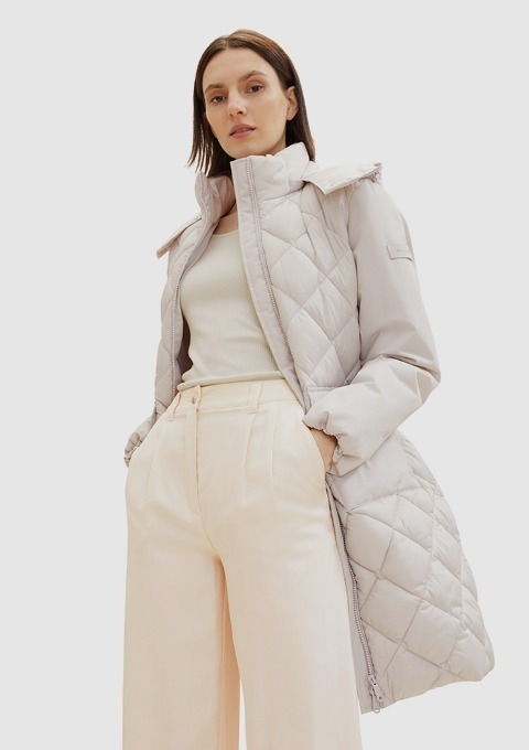 Eine Person trägt einen grauen, gepolsterten Mantel und eine hellbeige Hose, mit einem neutralen Hintergrund.