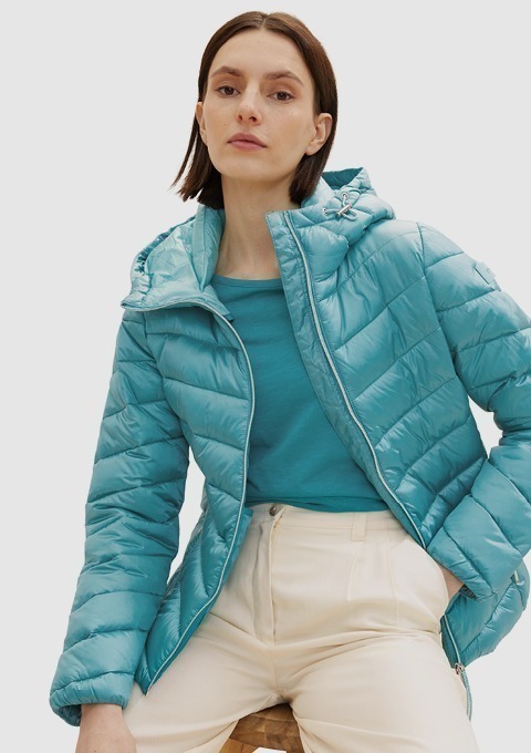 Eine Frau trägt eine türkisfarbene, gesteppten Jacke und ein blaues Oberteil. Sie sitzt auf einem Hocker und schaut direkt in die Kamera.