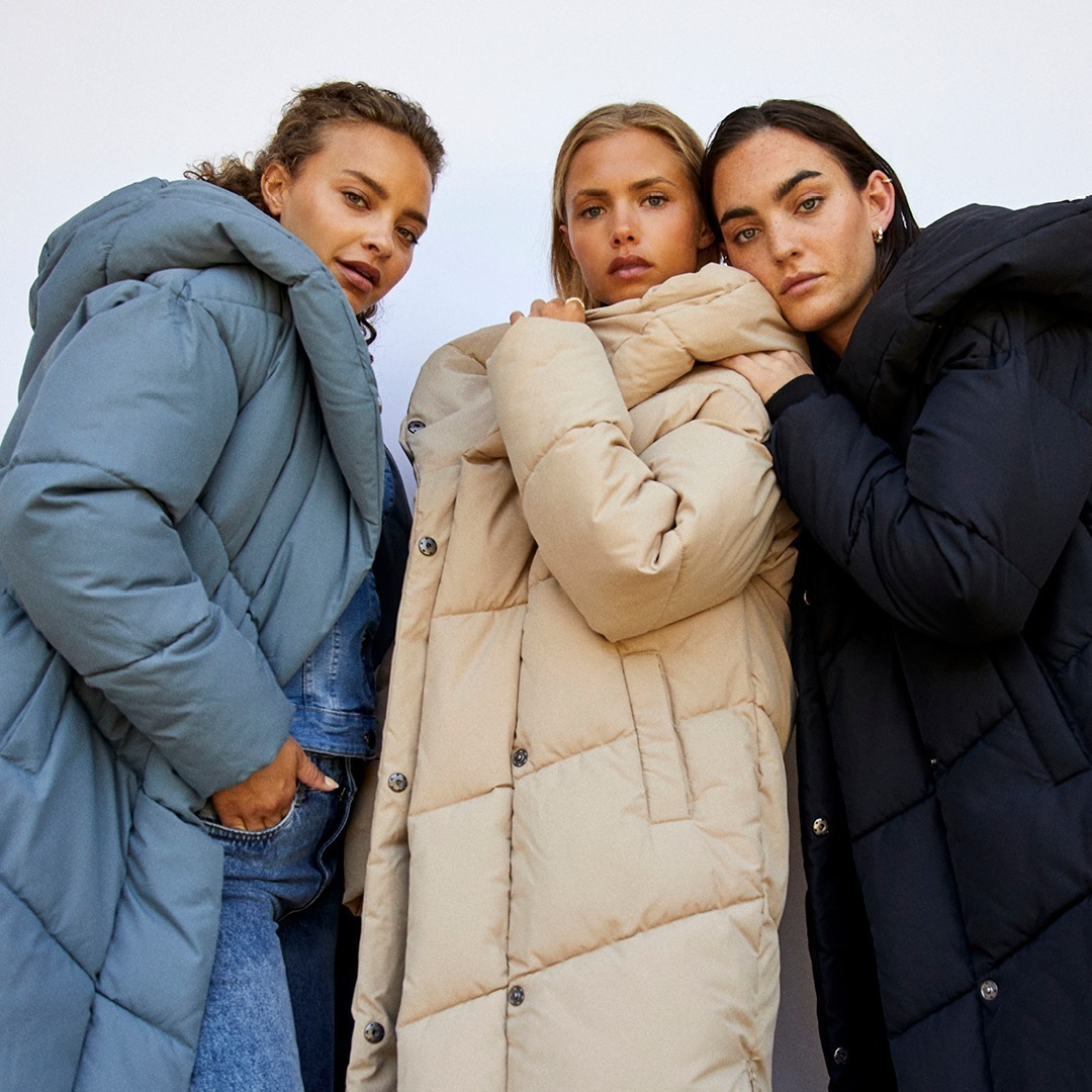 Drei Frauen stehen zusammen, jede trägt eine dicke, gepolsterte Jacke in verschiedenen Farben: blau, beige und schwarz.