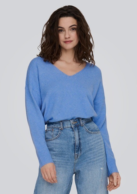 Eine Person mit dunklen, lockigen Haaren trägt einen blauen Pullover und eine Jeans.
