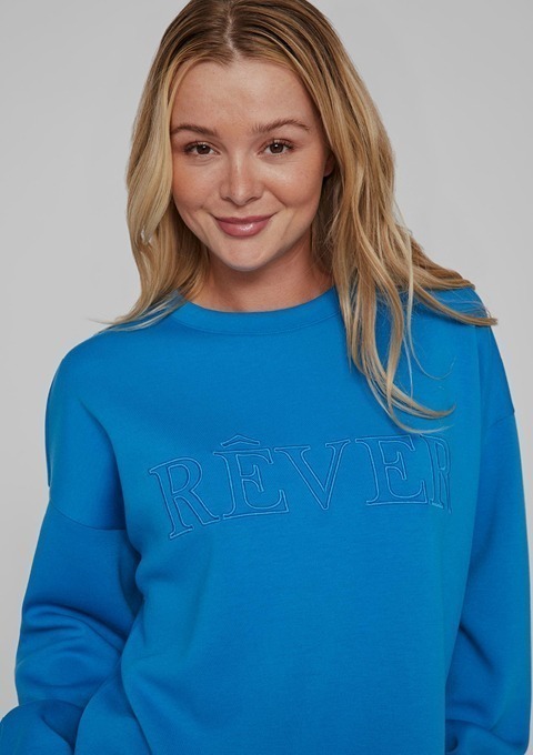 Eine lächelnde Frau trägt einen blauen Pullover mit der Aufschrift „RÊVER“.