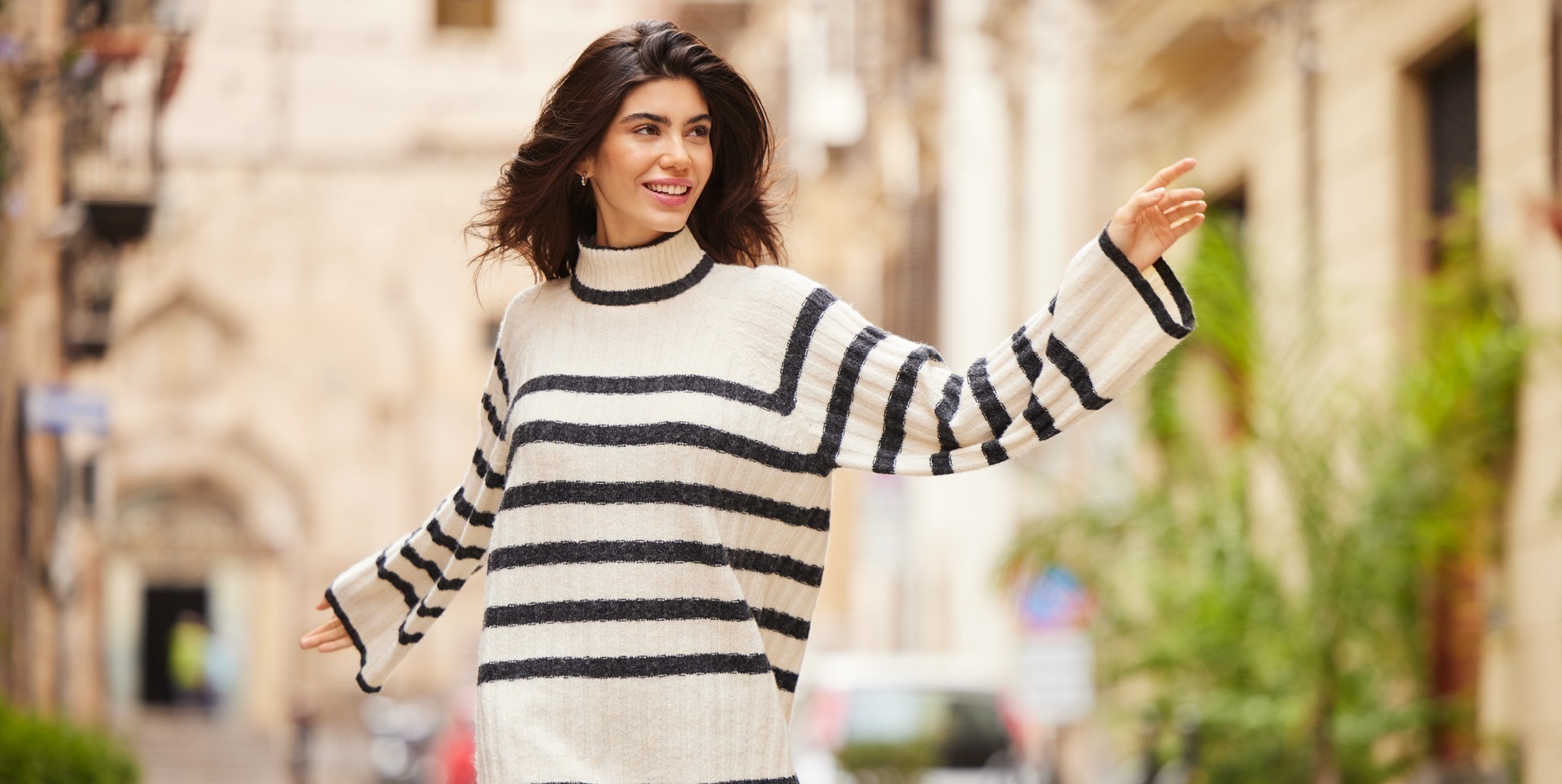 Eine Frau mit langen Haaren trägt einen gestreiften Pullover und lächelt, während sie in einer urbanen Straße posiert.