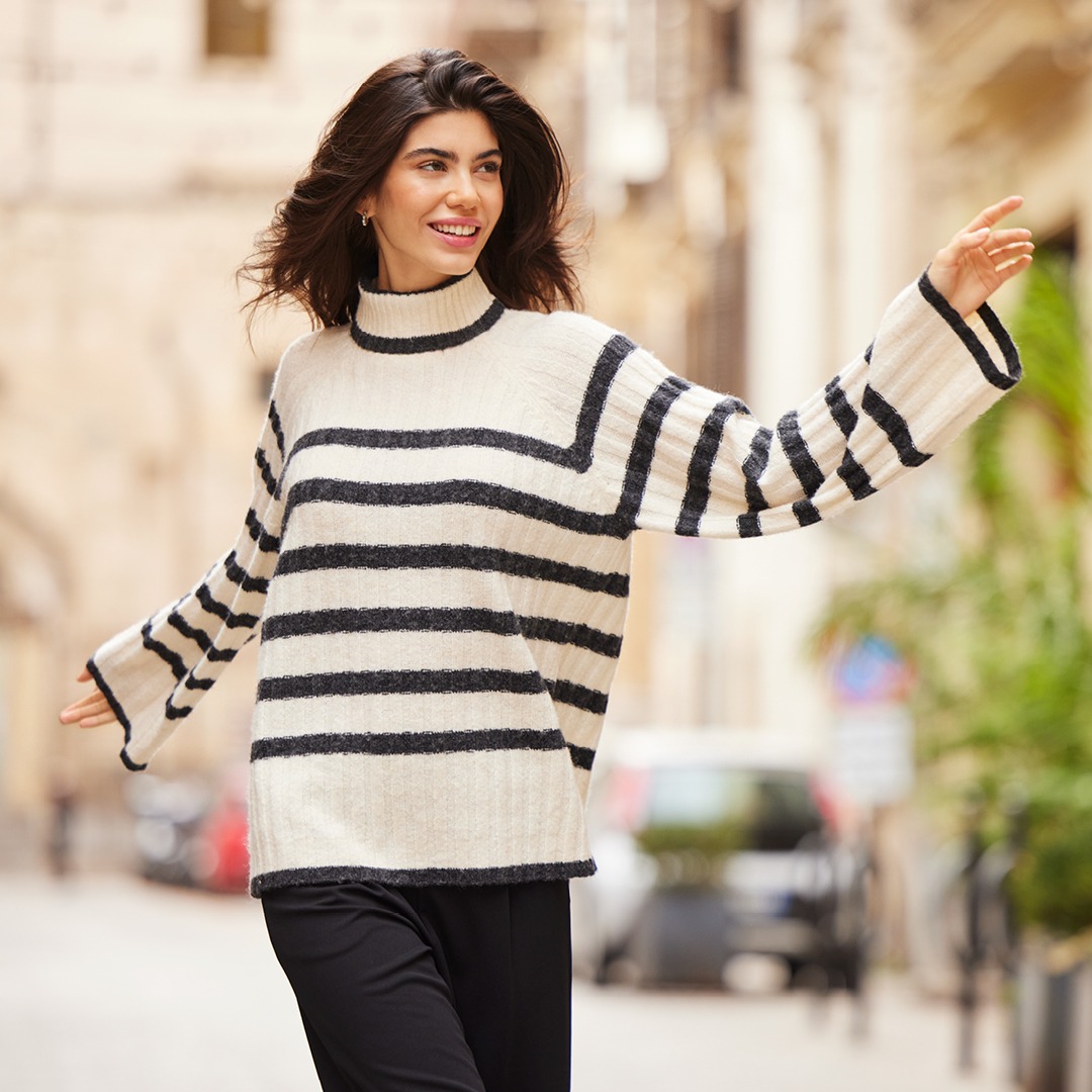 Eine Frau mit langen Haaren trägt einen gestreiften Pullover und steht in einer Stadtstraße, lächelnd und mit ausgebreiteten Armen. Eine Frau mit langen Haaren trägt einen gestreiften Pullover und steht in einer Stadtstraße, lächelnd und mit ausgebreiteten Armen.
