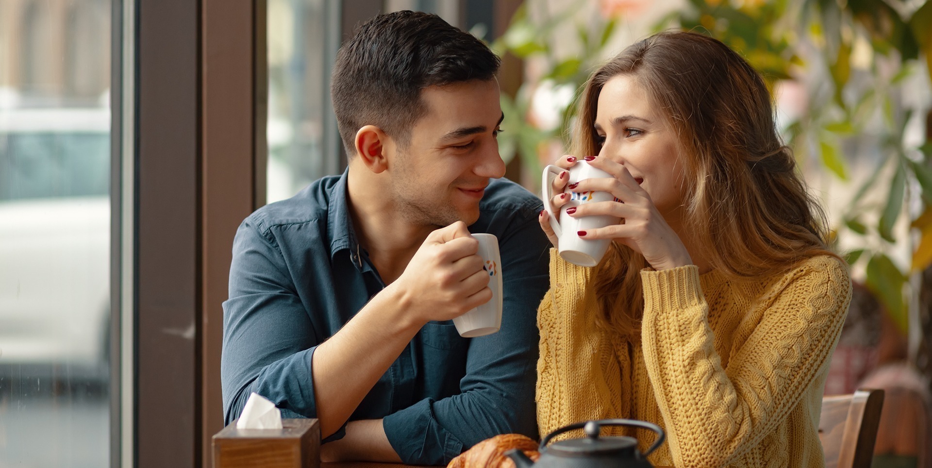 Ein Paar sitzt in einem Café und trinkt aus Kaffeetassen, während sie sich anlächeln.