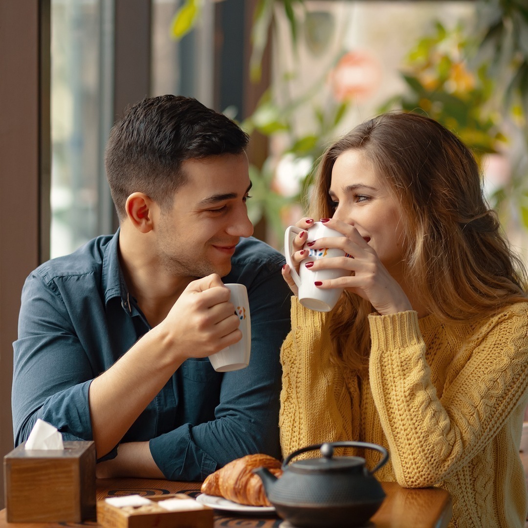 Ein Paar sitzt in einem Café, erhebt Tassen und lächelt sich an. Auf dem Tisch liegen ein Croissant und eine Teekanne.
