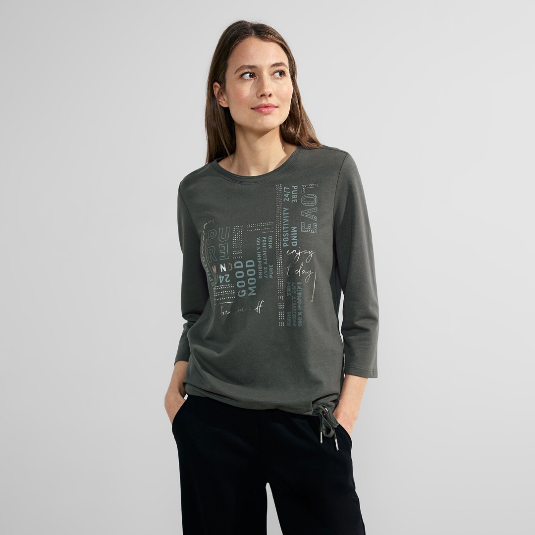 Eine Frau in einem grauen Langarmshirt mit Aufdruck steht vor einem neutralen Hintergrund. Eine Frau in einem grauen Langarmshirt mit Aufdruck steht vor einem neutralen Hintergrund.