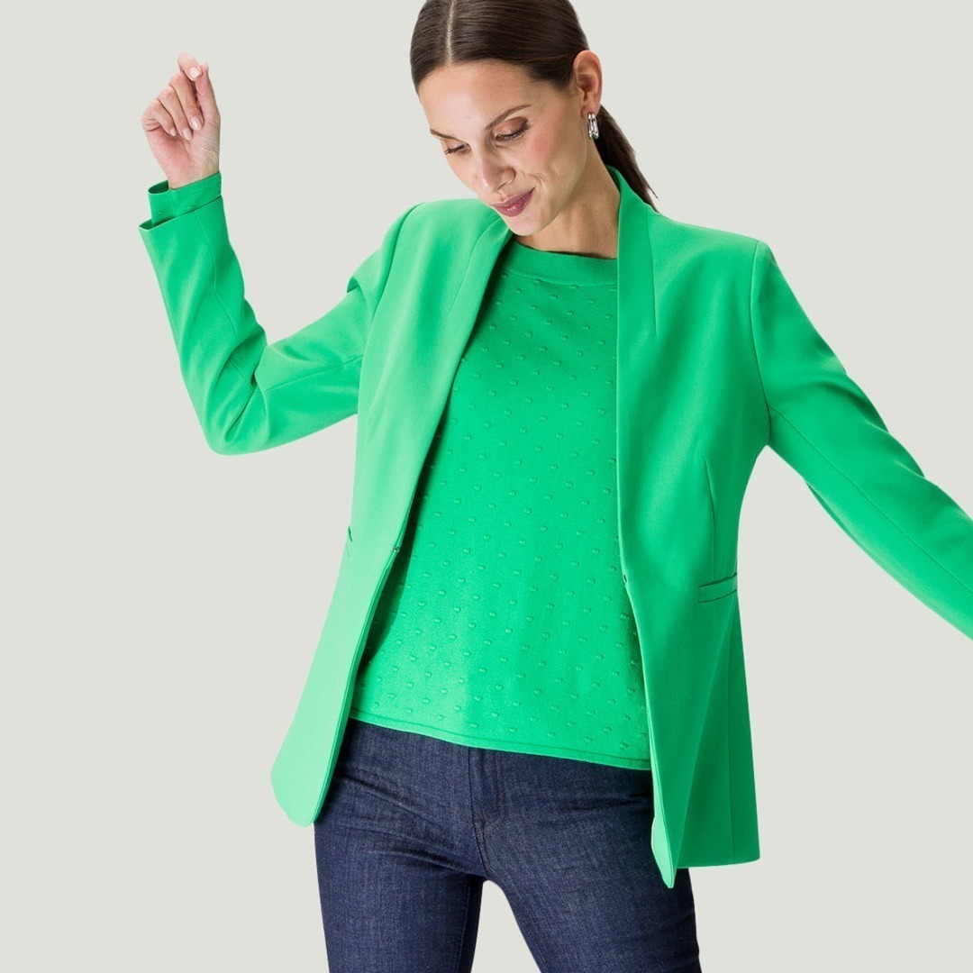Eine Frau trägt einen grünen Blazer und ein grünes Oberteil. Sie schaut lächelnd auf den Boden und hebt ein Arm. Eine Frau trägt einen grünen Blazer und ein grünes Oberteil. Sie schaut lächelnd auf den Boden und hebt ein Arm.