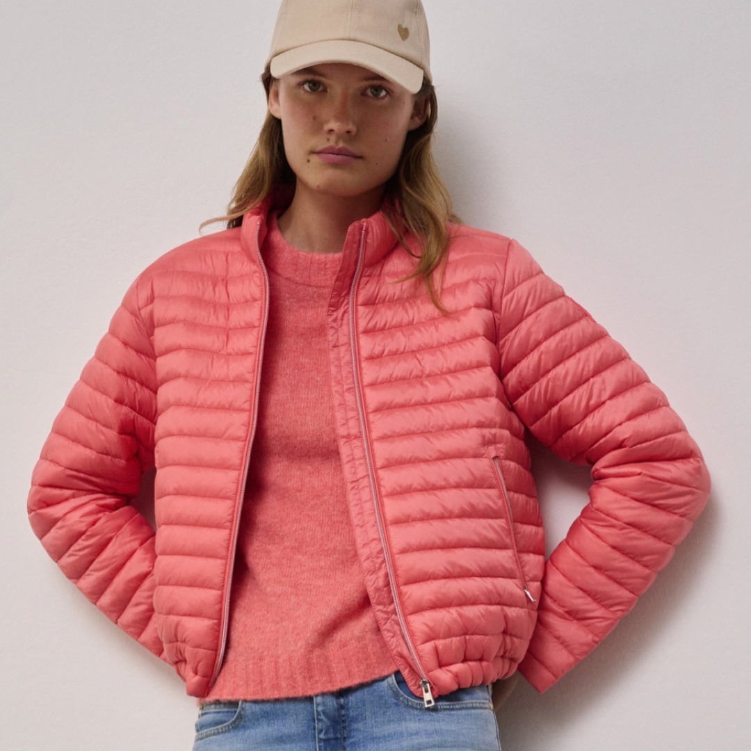 Eine Frau trägt eine rosafarbene, gesteppte Jacke und einen beigen Hut. Eine Frau trägt eine rosafarbene, gesteppte Jacke und einen beigen Hut.