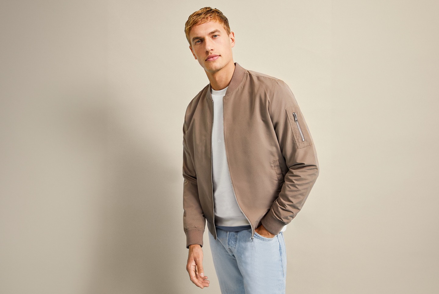 Ein Mann trägt eine beige Bomberjacke und eine weiße T-shirt, steht vor einem hellen Hintergrund. Ein Mann trägt eine beige Bomberjacke und eine weiße T-shirt, steht vor einem hellen Hintergrund.