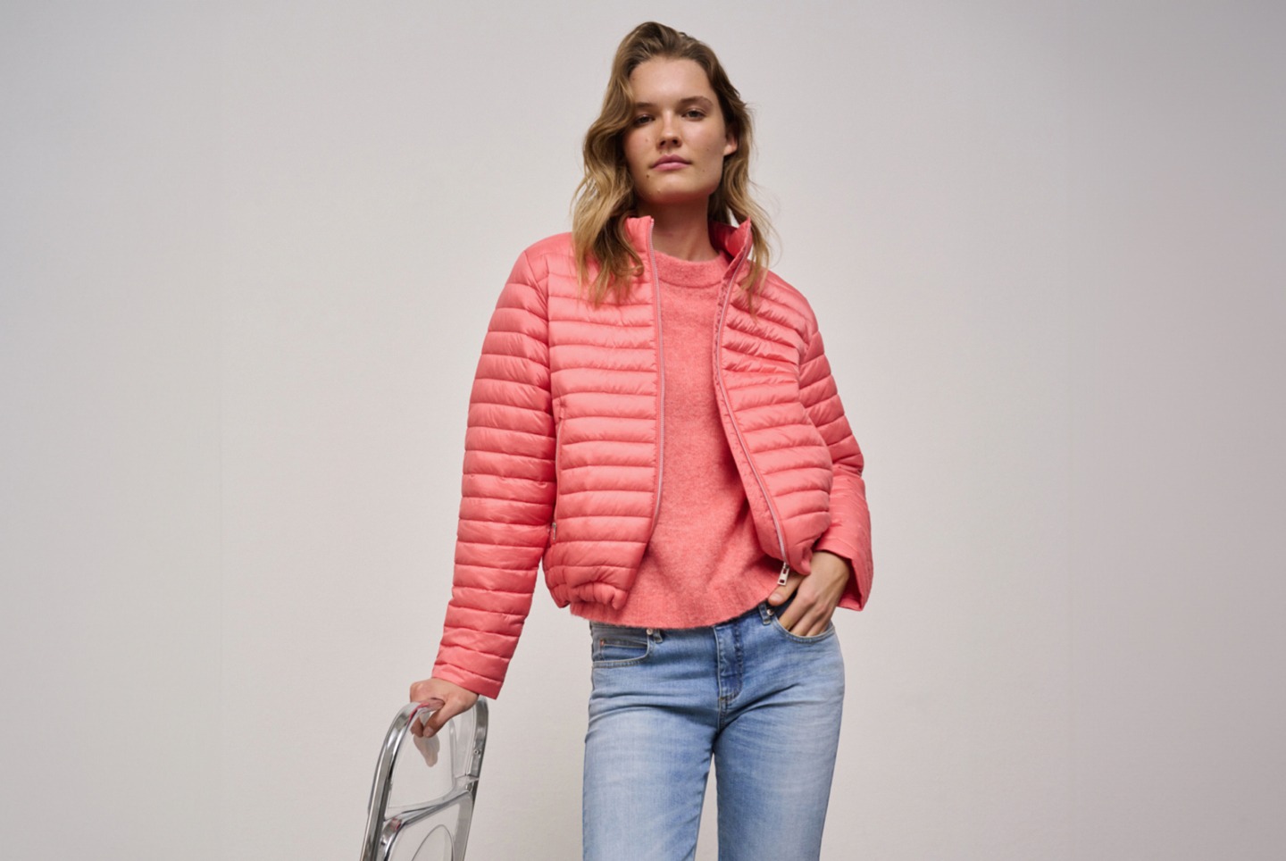 Eine Frau trägt eine rosa Steppjacke und ein passendes Oberteil, während sie eine durchsichtige Stuhllehne hält. Eine Frau trägt eine rosa Steppjacke und ein passendes Oberteil, während sie eine durchsichtige Stuhllehne hält.
