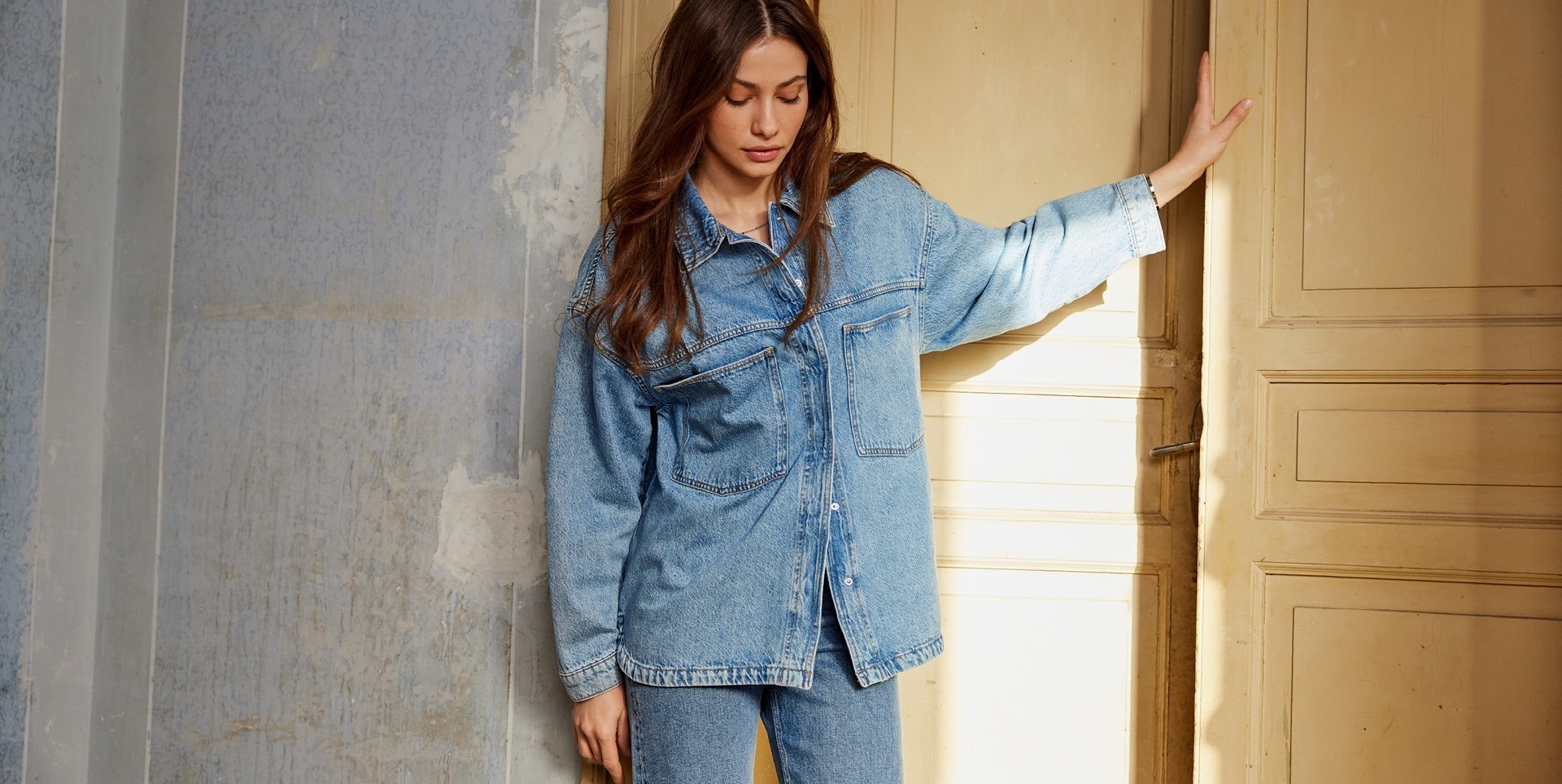 Eine Frau trägt eine Jeansjacke und Jeans. Sie steht in einem Raum mit blauen Wänden und einer offenen Tür.