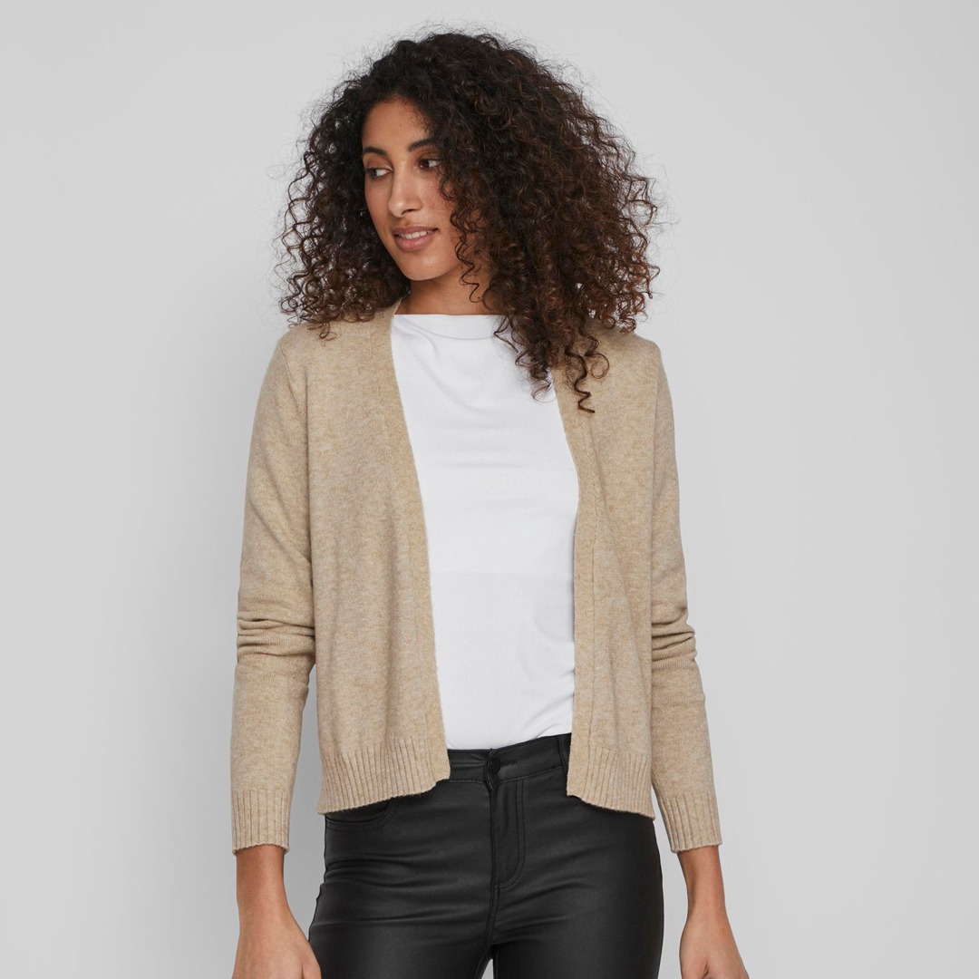 Eine Frau mit lockigem, brünettem Haar trägt einen beigen Cardigan über einem weißen T-Shirt und schwarzen Hosen. Eine Frau mit lockigem, brünettem Haar trägt einen beigen Cardigan über einem weißen T-Shirt und schwarzen Hosen.