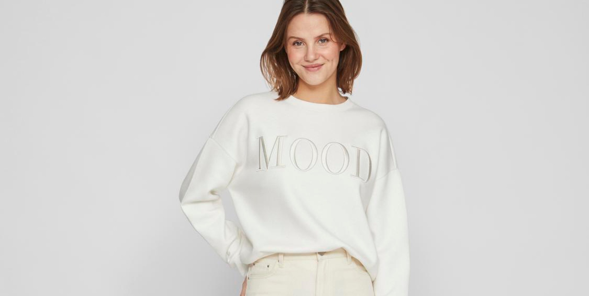 Eine lächelnde Frau trägt einen weißen Pullover mit der Aufschrift "MOOD" und helle Jeans.