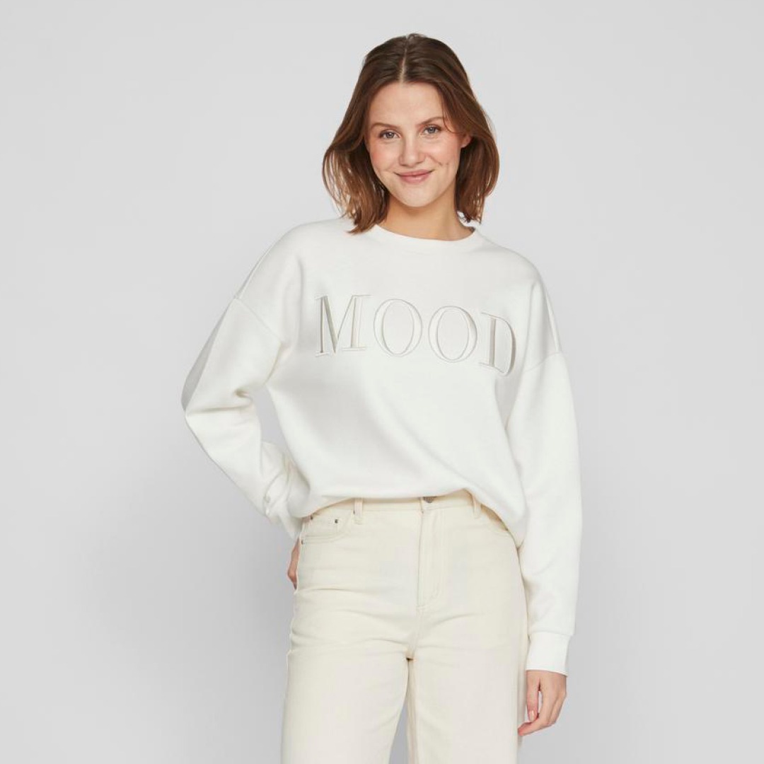 Eine Frau trägt einen weißen Sweatshirt mit dem Aufdruck „MOOD“ und helle Jeans.
