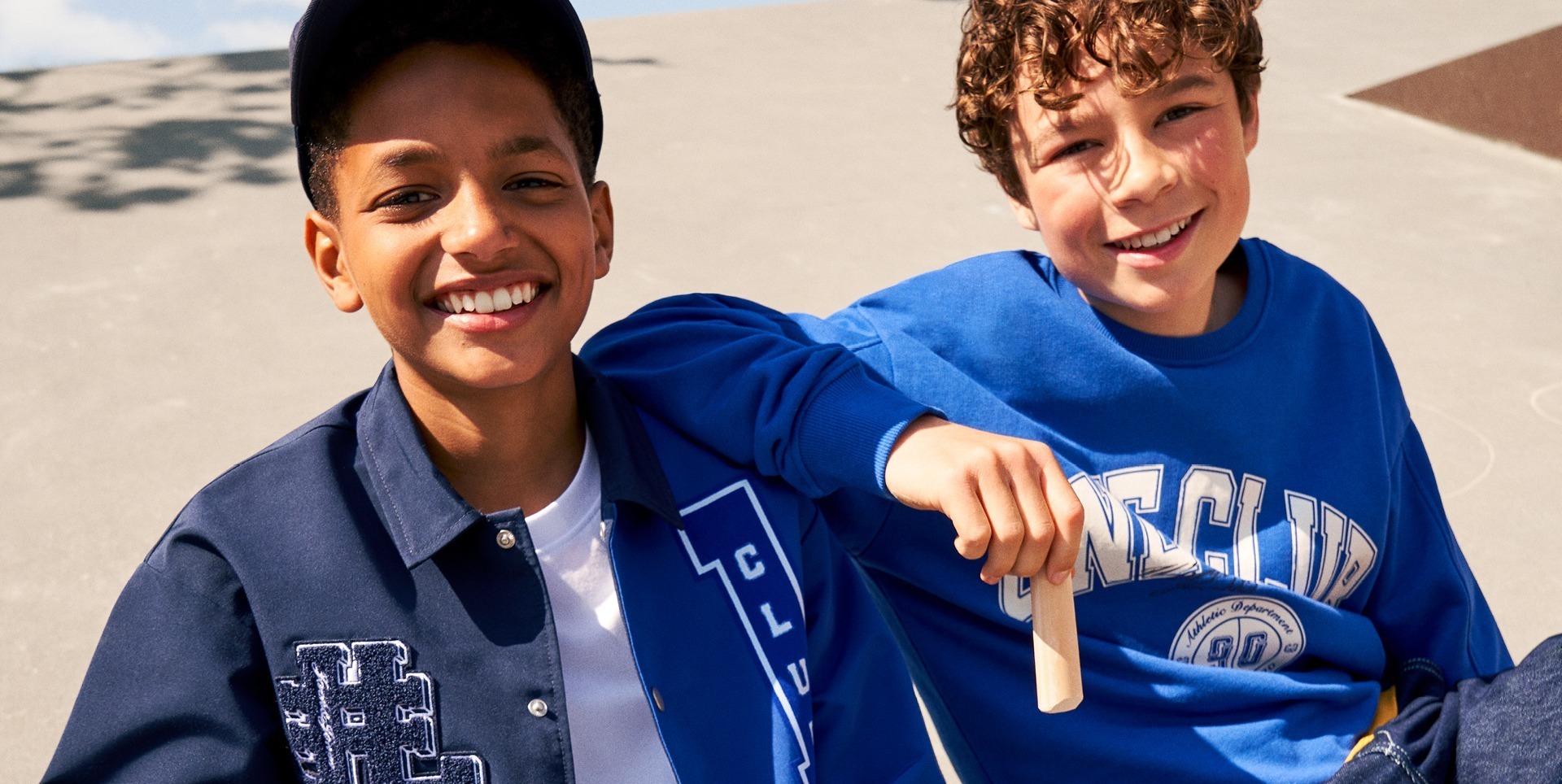 Zwei lächelnde Jungen, einer in einem blauen Sweatshirt und der andere in einer blauen Jacke, sitzen zusammen und genießen die Sonne.