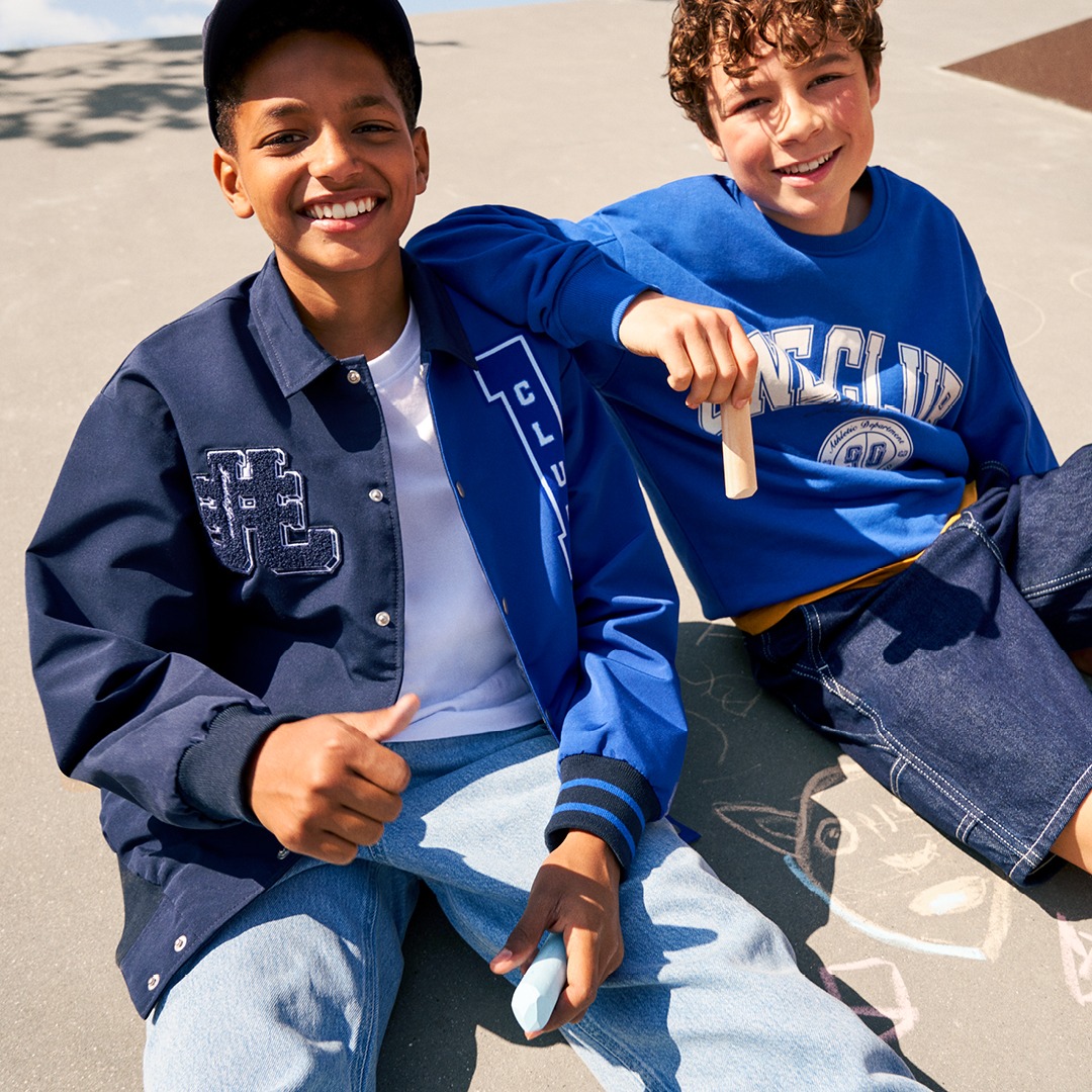 Zwei lächelnde Jungen sitzen auf einem befestigten Boden. Der Junge links trägt eine dunkelblaue Jacke, der rechte ein blaues Sweatshirt. Zwei lächelnde Jungen sitzen auf einem befestigten Boden. Der Junge links trägt eine dunkelblaue Jacke, der rechte ein blaues Sweatshirt.