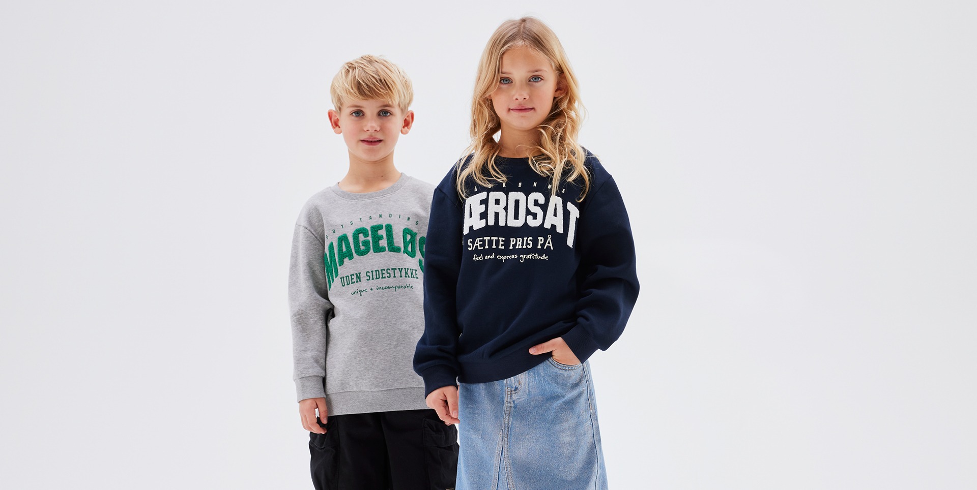 Ein Junge und ein Mädchen stehen nebeneinander. Der Junge trägt einen grauen Pullover mit grüner Schrift, das Mädchen einen dunkelblauen Pullover mit weißer Schrift und einen Jeansrock. Ein Junge und ein Mädchen stehen nebeneinander. Der Junge trägt einen grauen Pullover mit grüner Schrift, das Mädchen einen dunkelblauen Pullover mit weißer Schrift und einen Jeansrock.