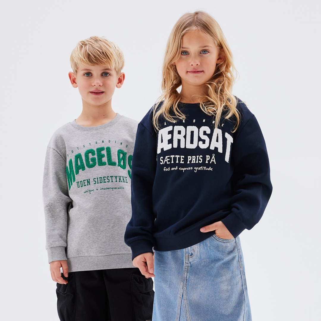 Zwei Kinder stehen nebeneinander, eines trägt einen grauen Pullover mit grüner Schrift, das andere ein dunkelblaues Sweatshirt. Zwei Kinder stehen nebeneinander, eines trägt einen grauen Pullover mit grüner Schrift, das andere ein dunkelblaues Sweatshirt.