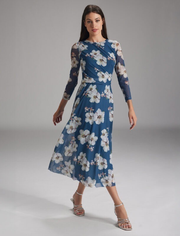 Eine Frau in einem blauen, blumigen Kleid mit langen Ärmeln, die auf einem neutralen Hintergrund steht.